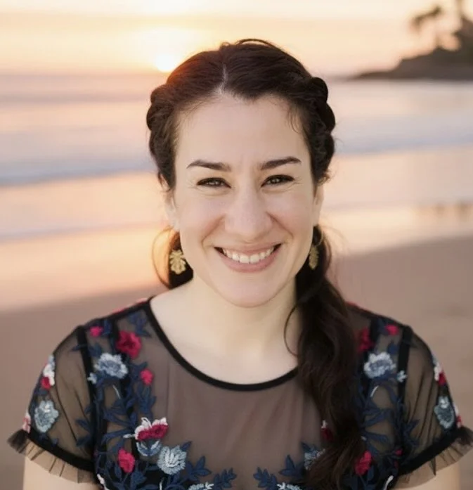 A woman with a braid wearing a black sheer top with colorful floral embroidery, smiling on a beach at sunset.