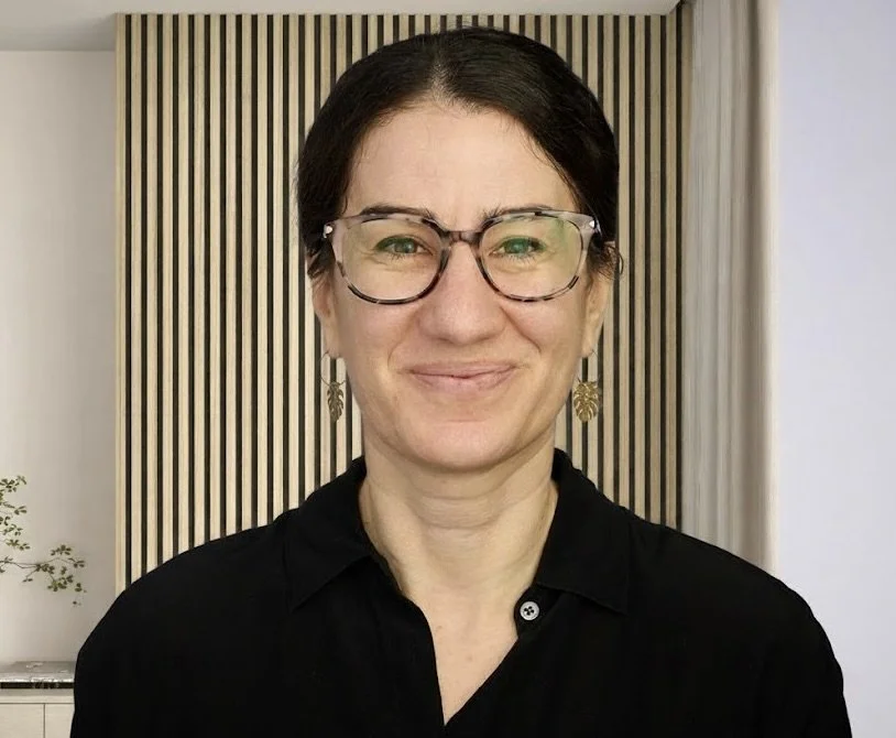 A woman with short dark hair, glasses, and earrings, smiling at the camera, standing in front of a modern wooden slat wall.