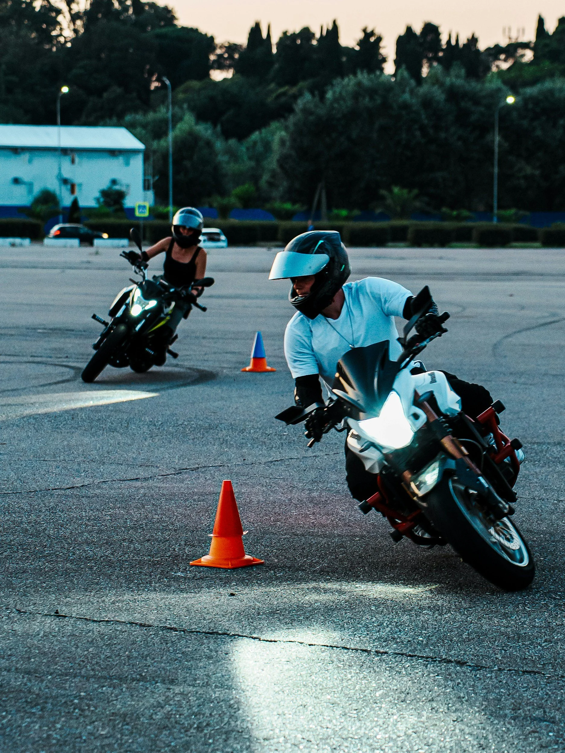 Two motorcyclists practicing riding skills on an empty parking lot, maneuvering around orange cones during dusk.