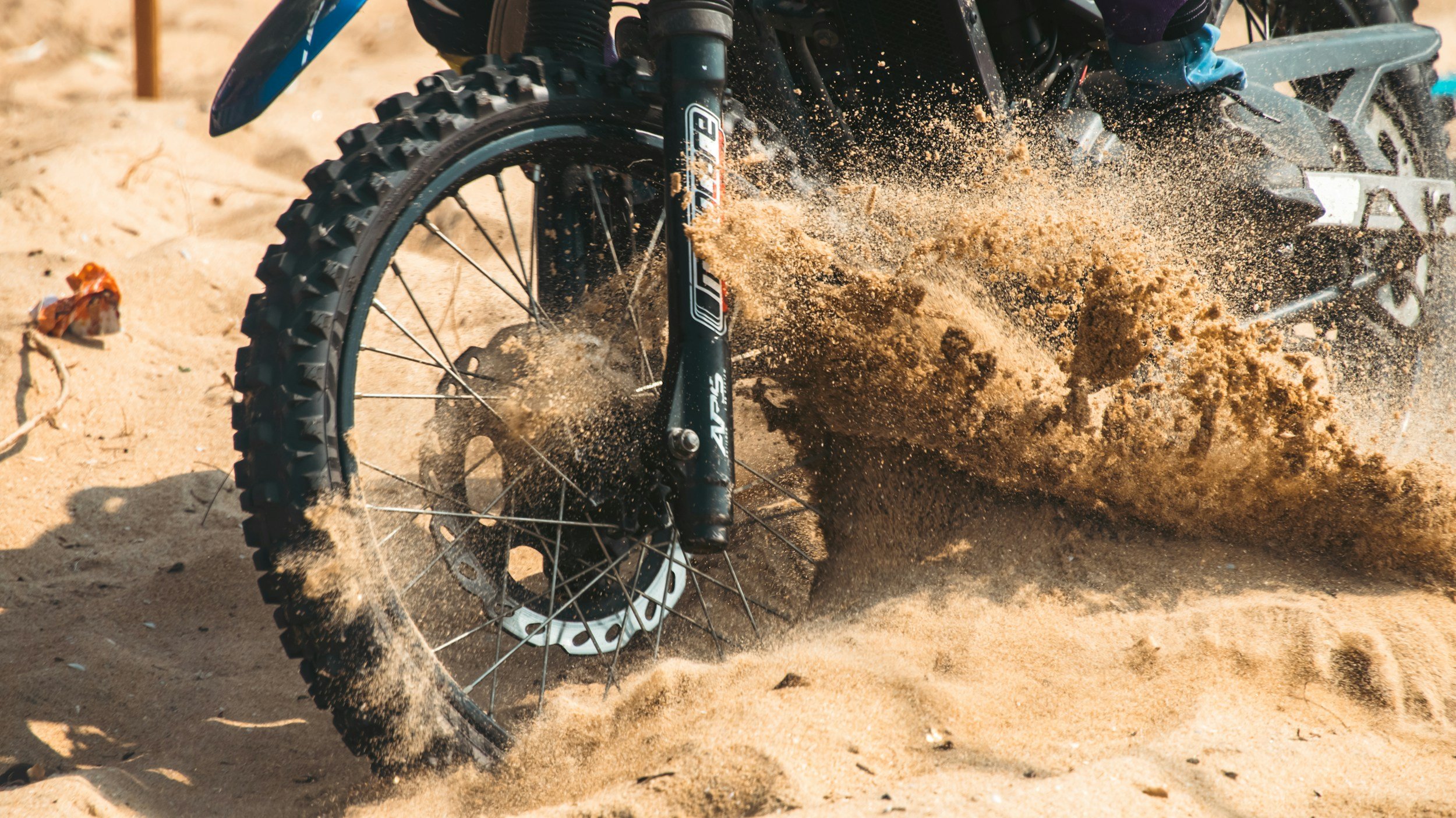 Close-up of a dirt bike tire kicking up sand on a sandy terrain, with visible sand flying in the air.