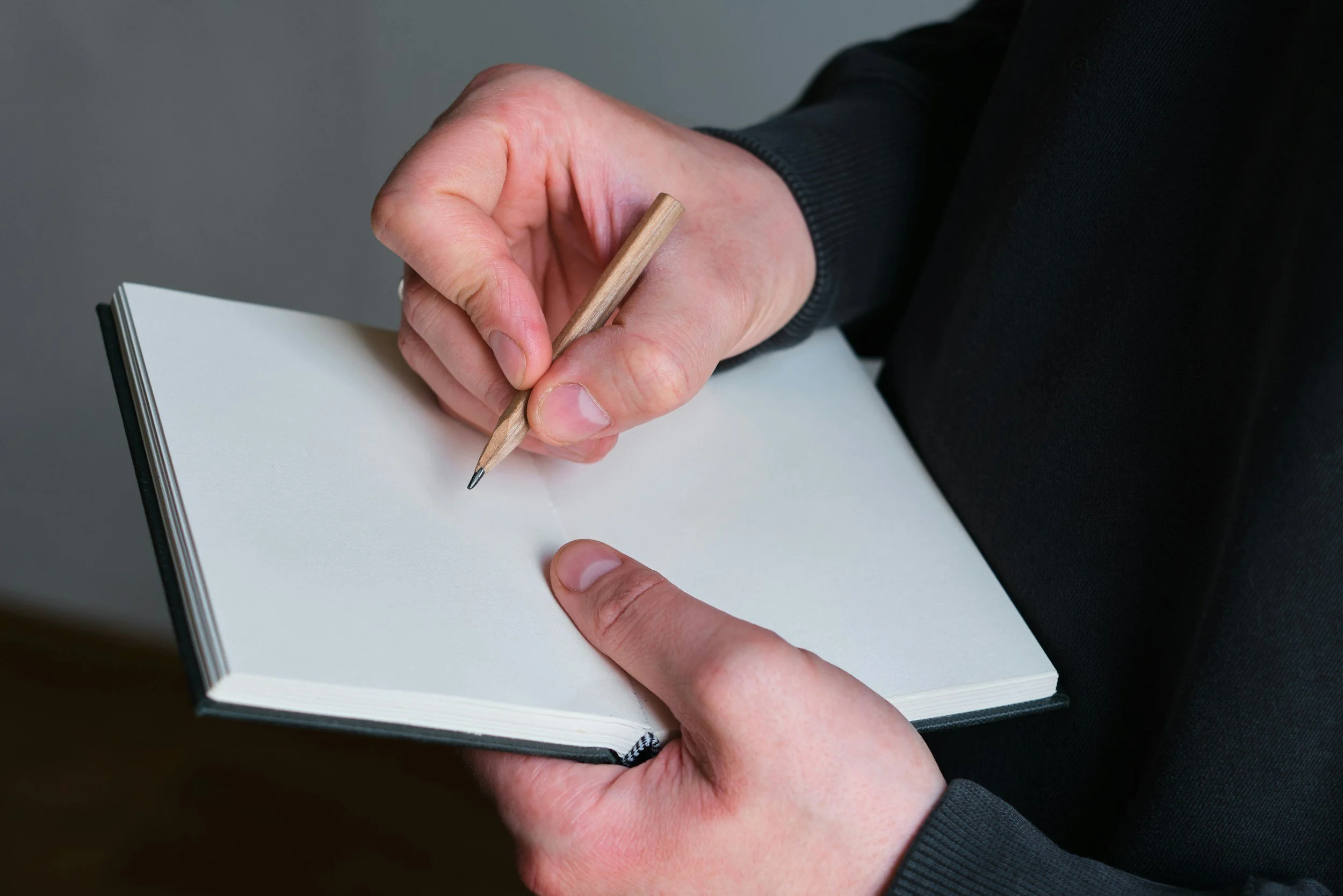 A person holding a small notepad and writing with a wooden pencil.