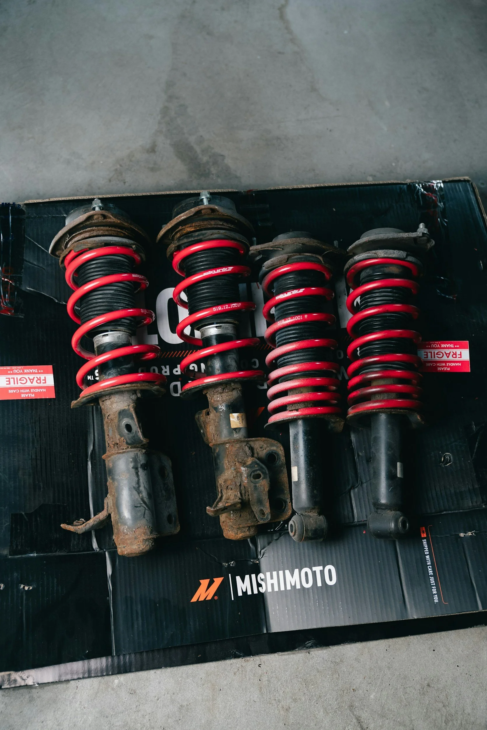 Four used motorcycle suspension shocks with red coil springs, placed on a black Mishimoto box, on a concrete floor.