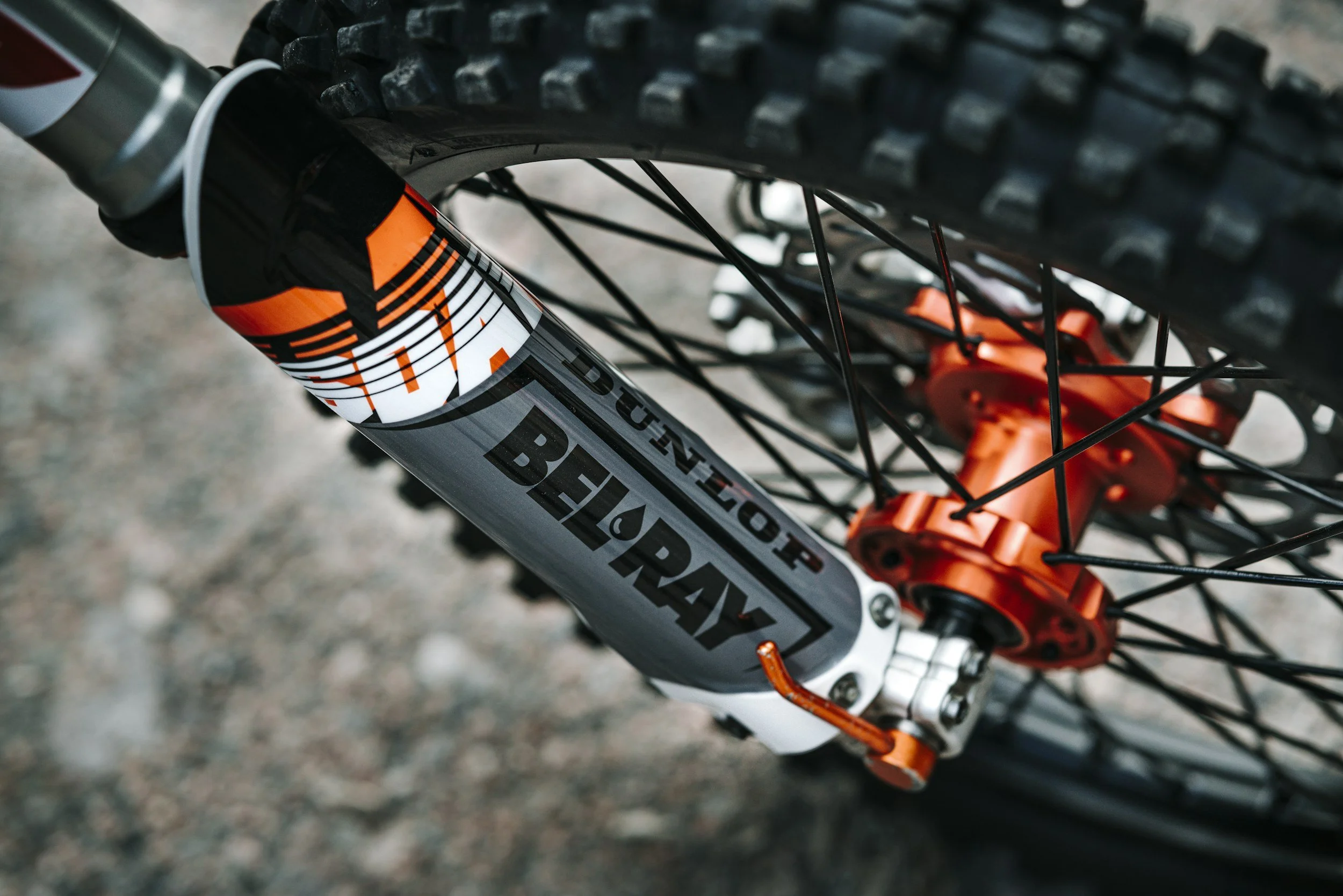 Close-up of a Motocross bike front suspension fork and wheel, with black spokes and an orange hub, on a gravel surface.