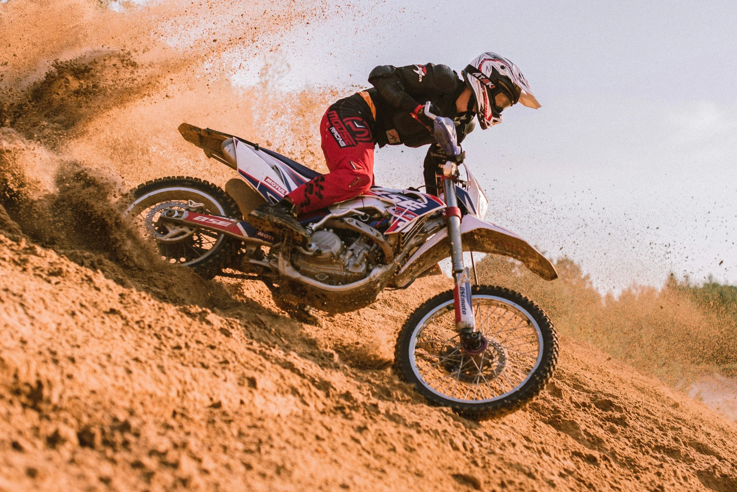 Motocross rider wearing protective gear and helmet riding on dirt track, kicking up dust.