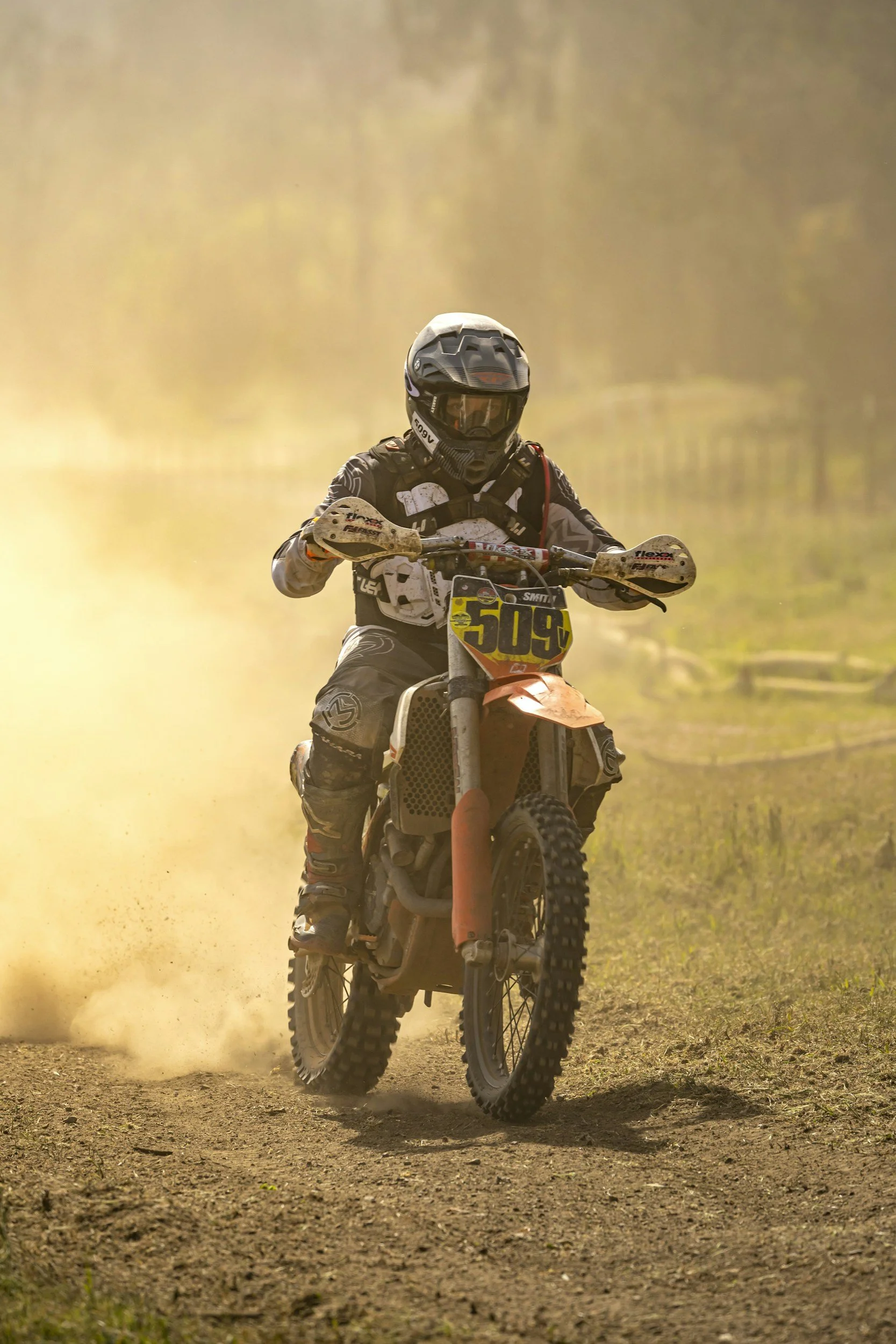 A dirt bike rider wearing protective gear driving on a dirt track with dust in the air, and a rural background.