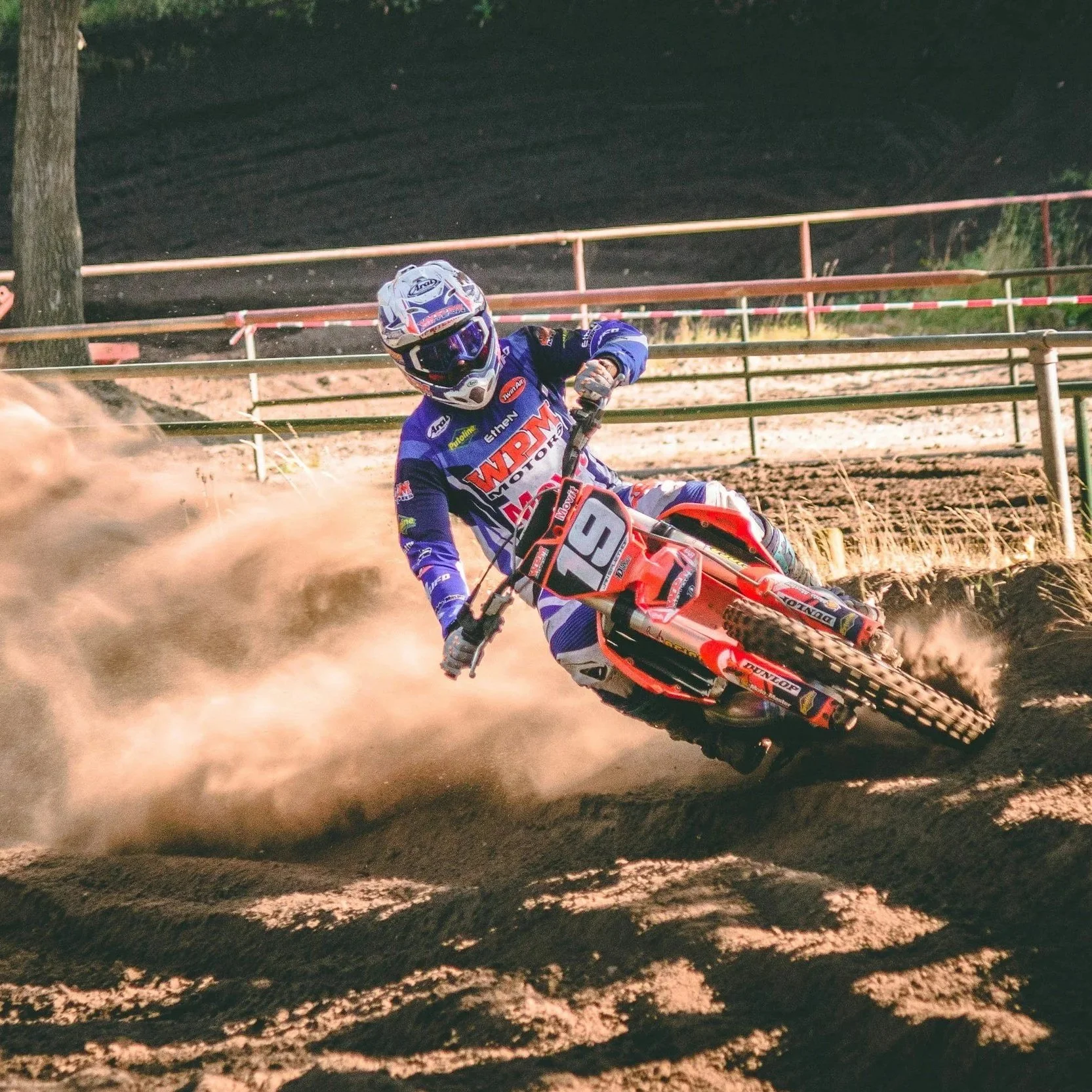 Motocross rider leaning into a turn on a dirt track, kicking up dust, wearing full protective gear and a blue racing suit, with a red and black dirt bike numbered 19.