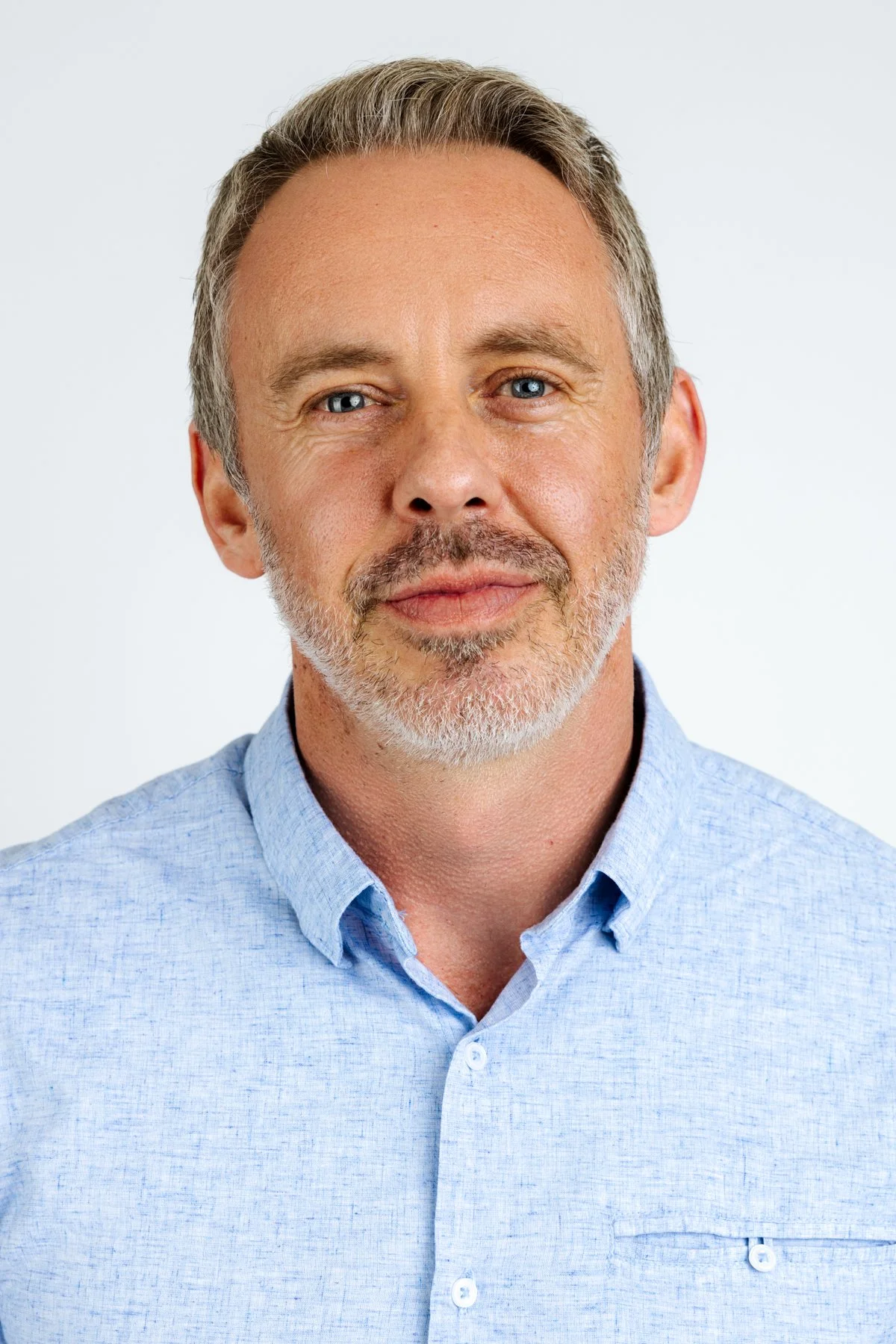 Dr Reid Robison, Chief Medical Officer. Portrait of a middle-aged man with gray hair, beard, and blue eyes, wearing a light blue button-up shirt, standing against a plain white background.