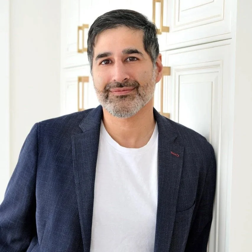 A middle-aged man with dark hair and a beard smiling, wearing a navy blazer over a white T-shirt, standing indoors against a white cabinet with gold handles.