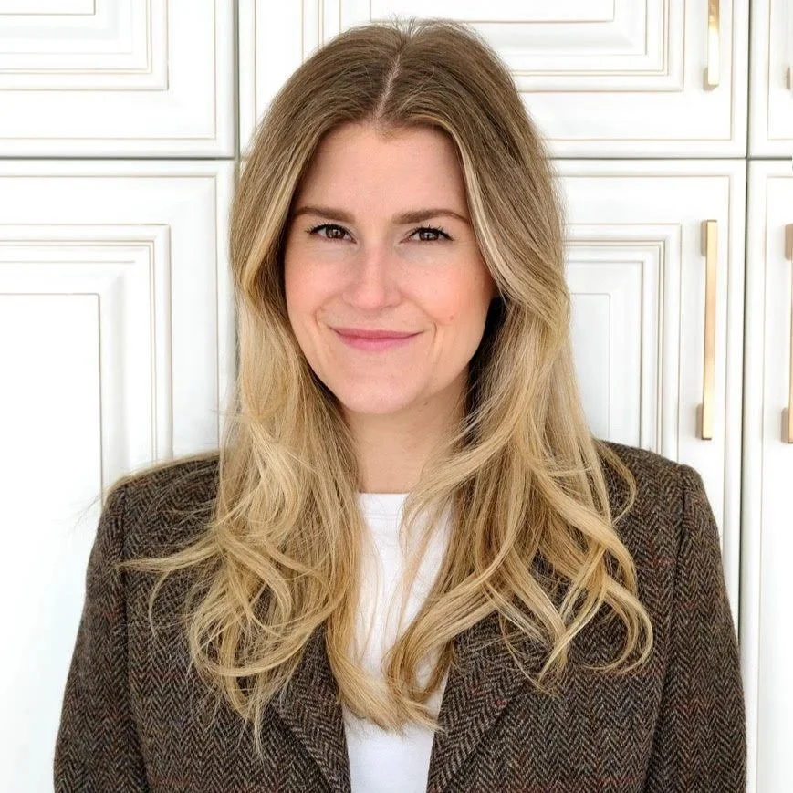 A woman with long blonde hair wearing a brown blazer with a white shirt, standing in front of white cabinets.