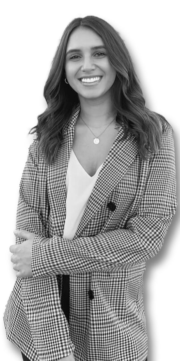 A woman with long wavy hair and a bright smile wearing a checkered blazer over a white top, standing outdoors with a blurred background.