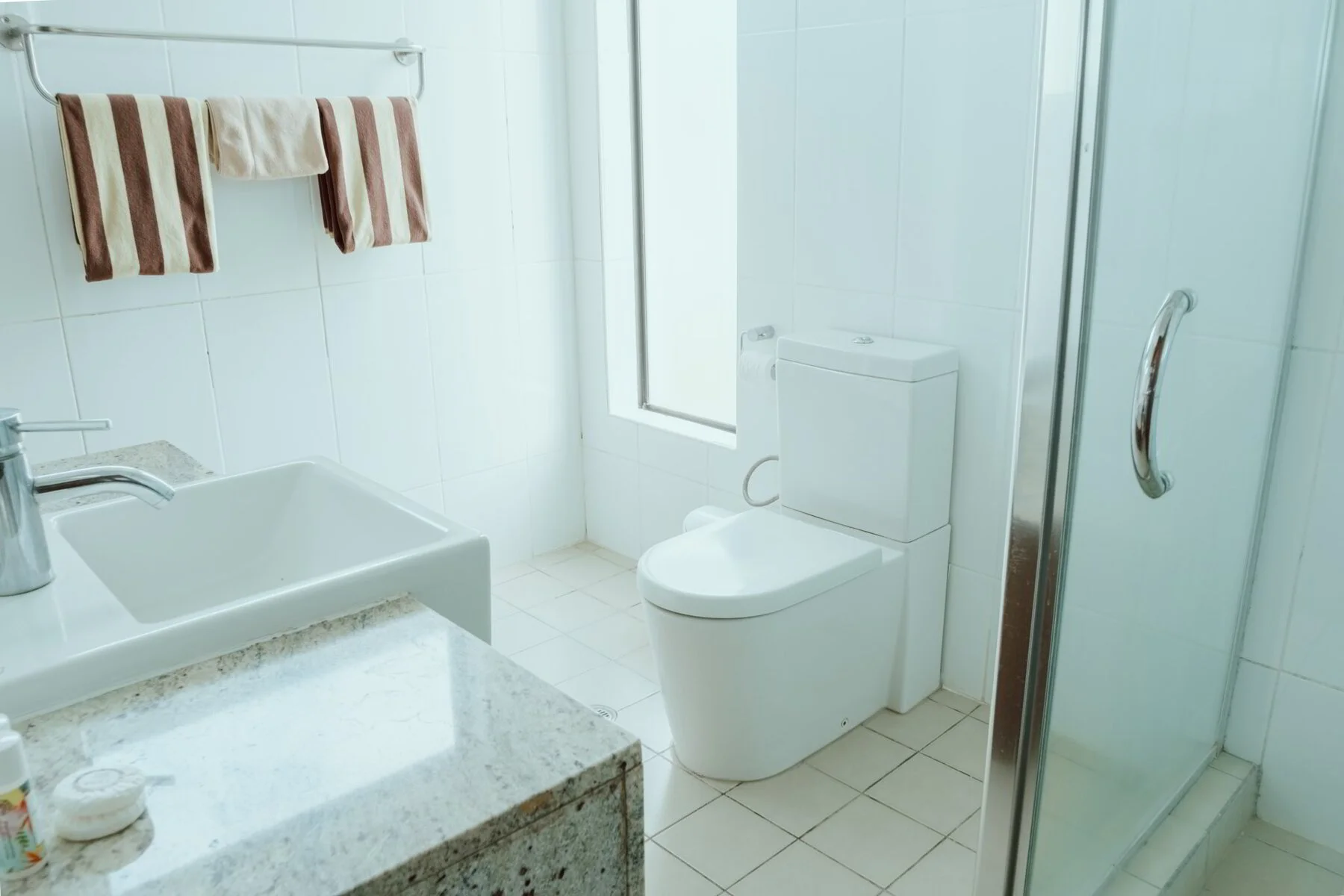 A clean bathroom with white tiled walls and floor, a white toilet, a small sink with a granite countertop, and striped towels hanging on a silver towel bar.