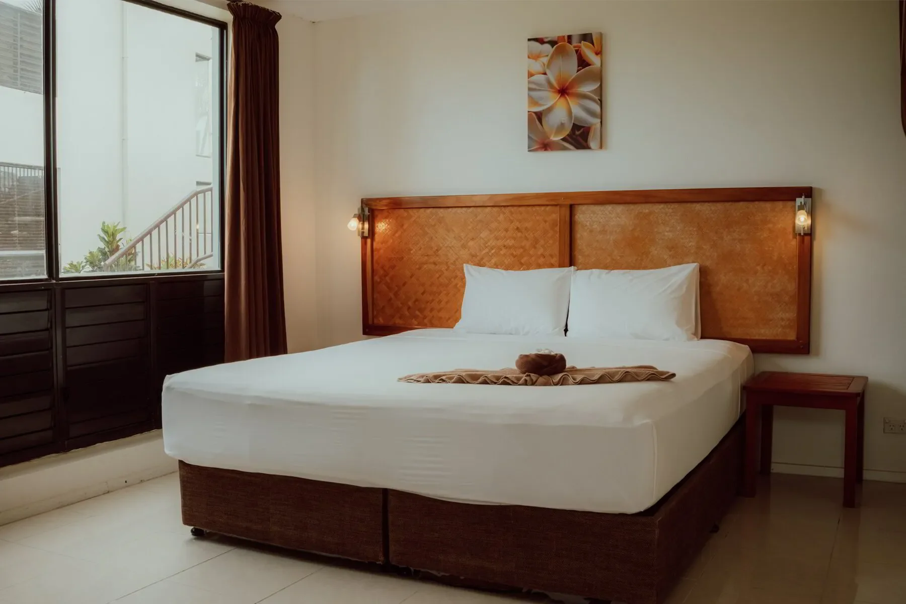 A hotel room with a large bed, white linens, and a wooden headboard, next to a window with brown curtains, and a nightstand on the right. There is a painting of flowers hanging above the bed.