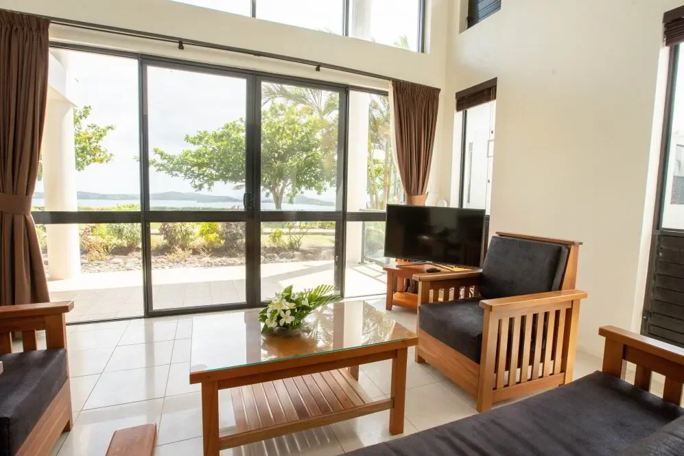 Living room with large sliding glass door overlooking an outdoor patio and greenery, furnished with wooden chairs, a coffee table with a flower arrangement, and a television.