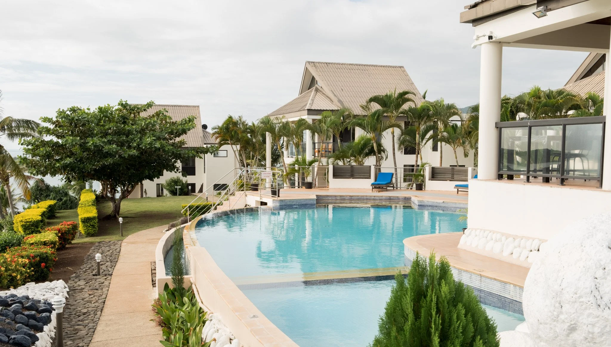 View of a tropical resort with a freeform swimming pool, surrounded by palm trees, colorful flowers, and white modern buildings with beige roofs.
