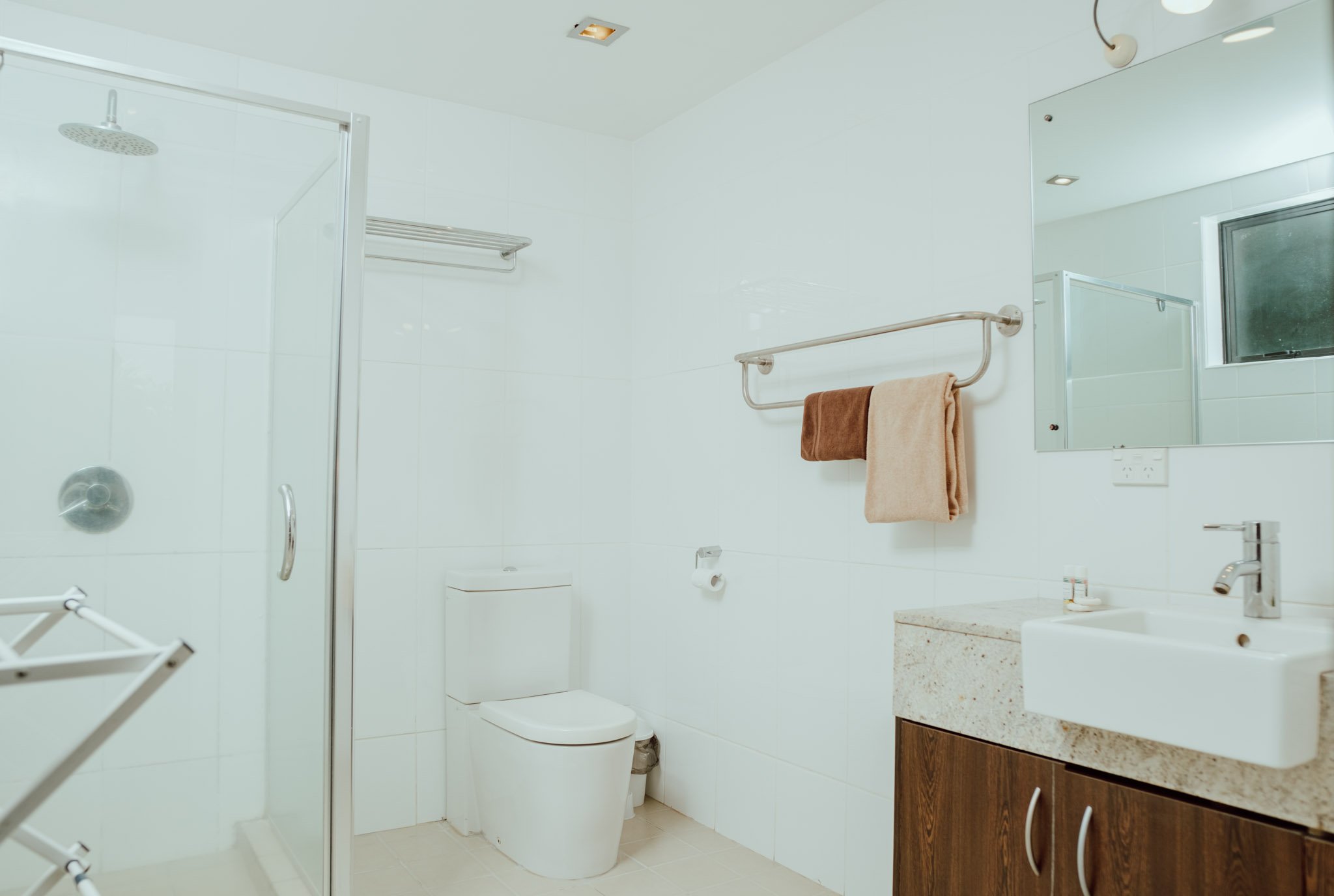 A clean, modern bathroom featuring a glass shower enclosure, a toilet, a small vanity with a sink, a mirror, and wall-mounted towel racks with towels. The bathroom has white tiles and a window for natural light.