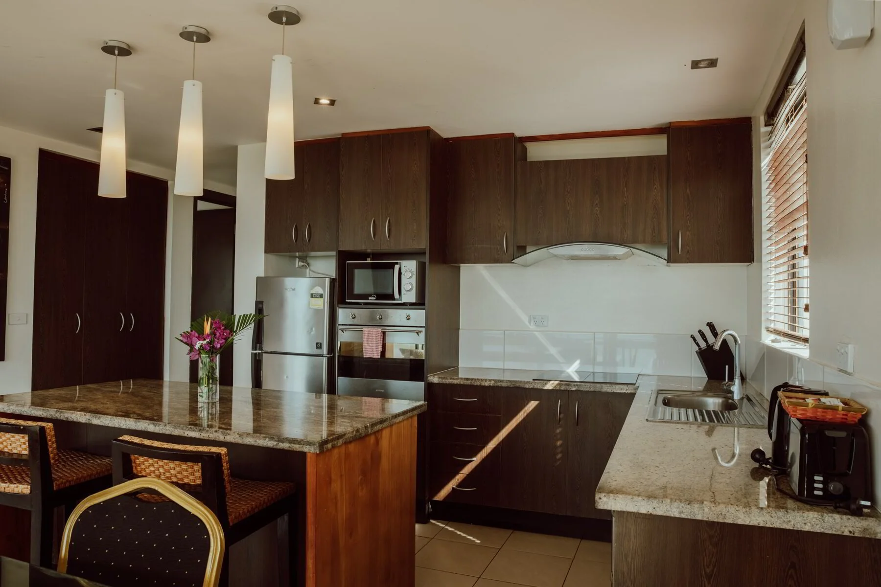 Modern kitchen with dark wooden cabinets, granite countertops, stainless steel appliances, and a small island with a vase of pink flowers.