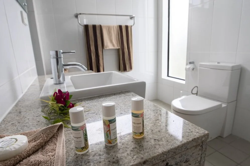 Bathroom with a granite countertop, a white sink, a chrome faucet, three small bottles of toiletries, a folded towel, a soap bar, a flower, a towel rack with two towels, a window, and a toilet.