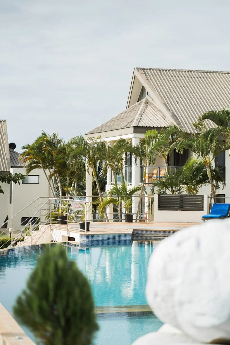 A house with a grey sloped roof, surrounded by palm trees, with a swimming pool in the foreground and a blue lounge chair on the poolside deck.