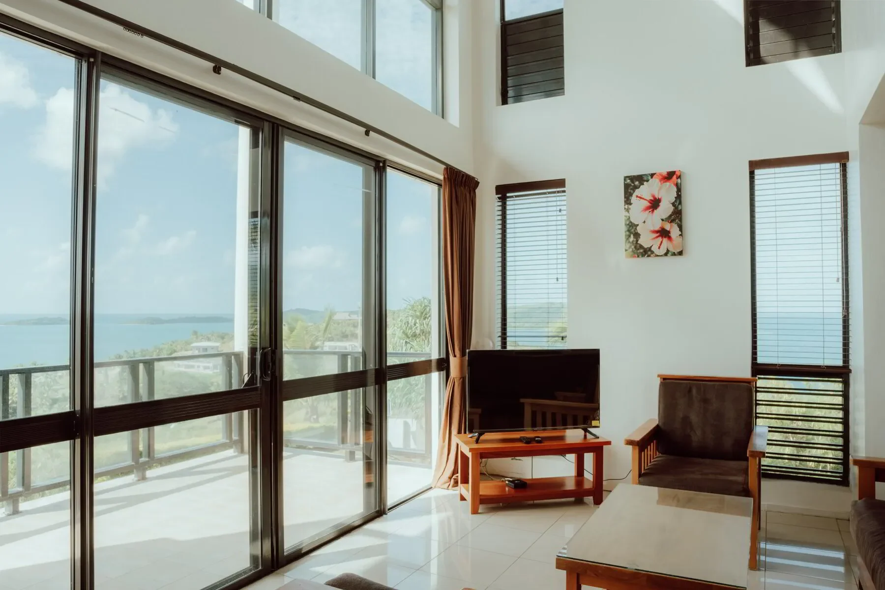 Living room with large sliding glass doors leading to a balcony, a flat-screen TV on a wooden stand, a wooden armchair, a small coffee table, and windows with blinds, overlooking a body of water and green landscape.