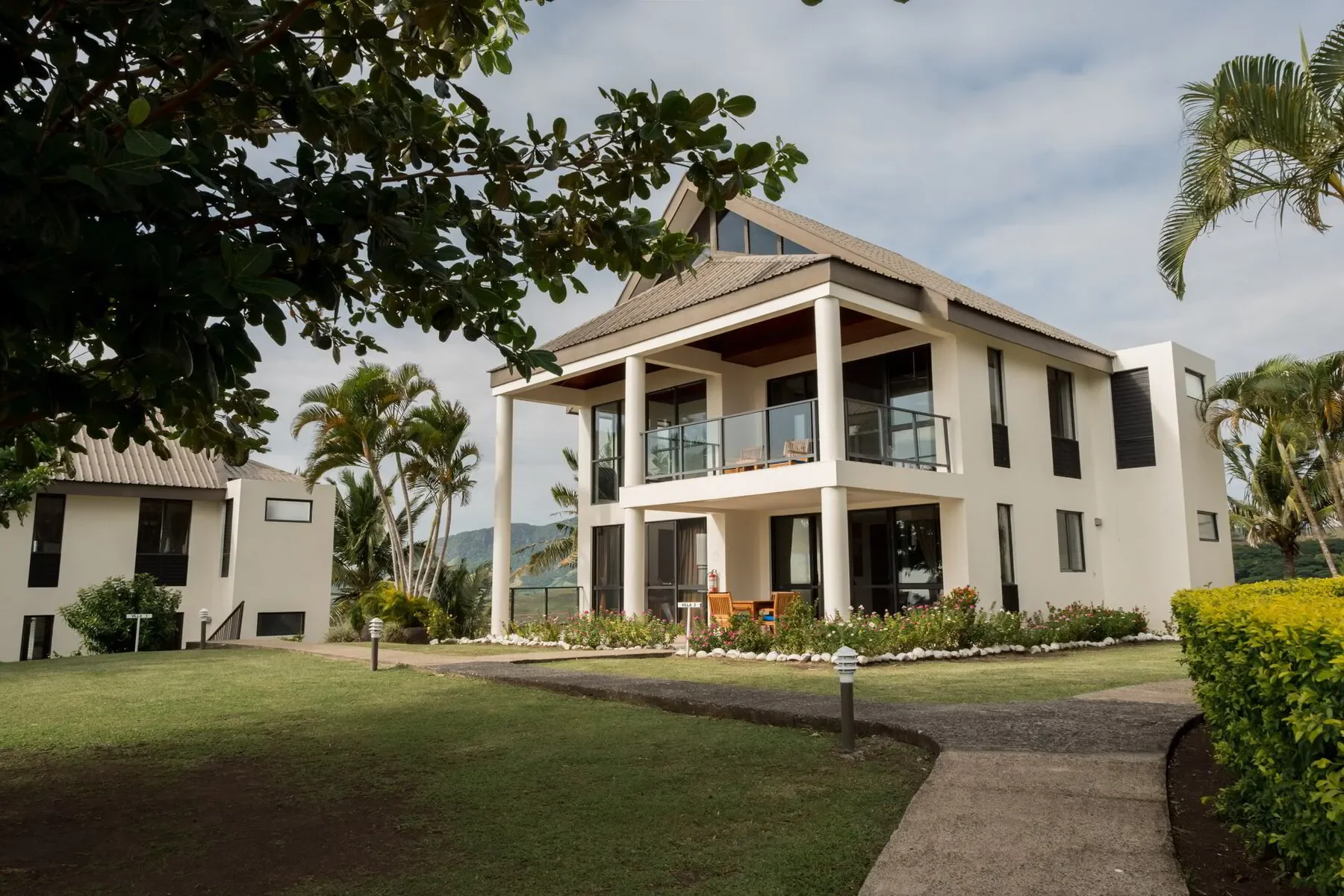 A modern two-story house with white exterior walls, large glass windows, and a balcony on the second floor, surrounded by a well-maintained lawn, bushes, and palm trees, under a partly cloudy sky.