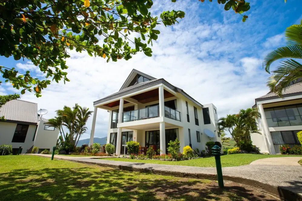 White modern house with glass balcony, surrounded by greenery and palm trees, under a blue sky with clouds.