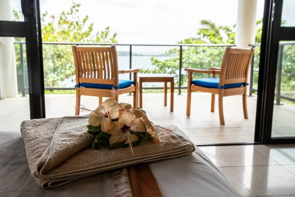 A view from inside a room looking out to a balcony with two wooden chairs with blue cushions and a small table, overlooking a body of water and green trees, with a towel and flower arrangement on a bed in the foreground.