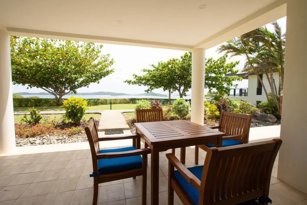 Covered patio with wooden table and four chairs, overlooking a garden with trees and a view of water in the distance.