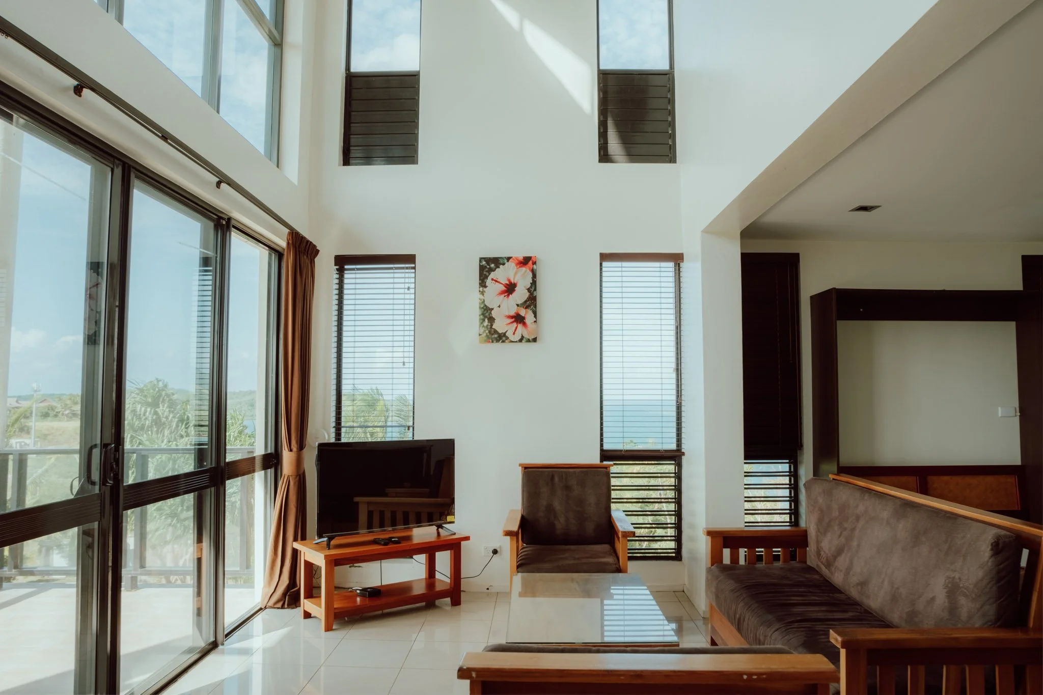 Interior view of a living room with large windows, a TV on a wooden stand, a dark sofa, a single armchair, and a glass-topped coffee table.