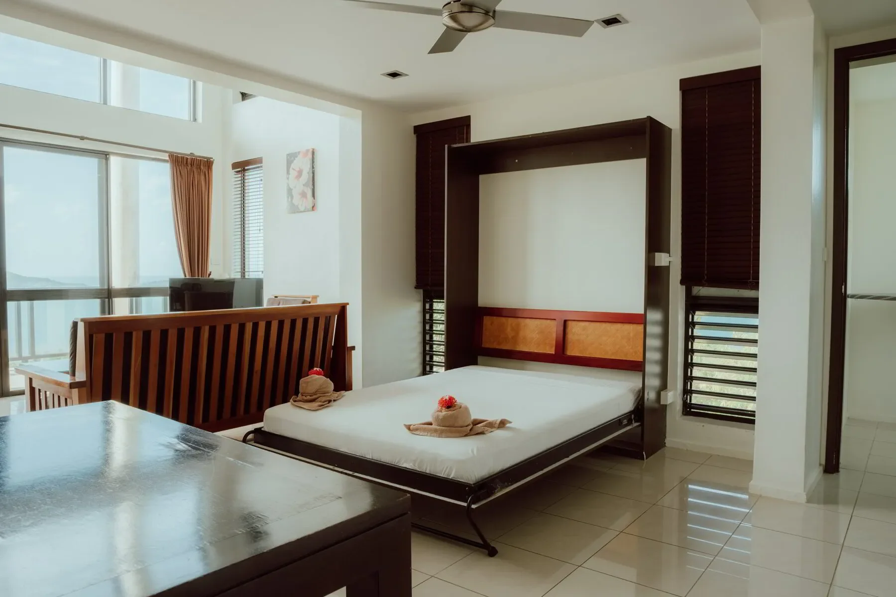 Hotel room with a bed that has towels folded into swan shapes with flowers on top, wooden furniture, windows with blinds, and a balcony view.