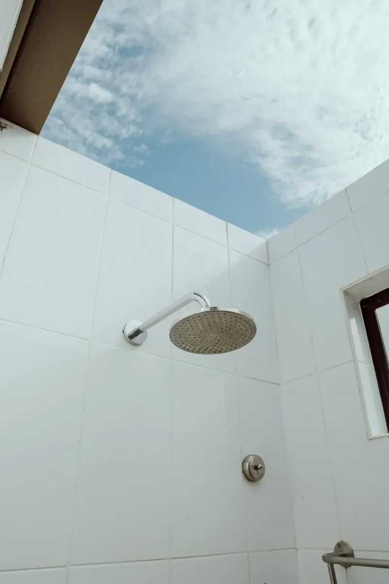 Showerhead mounted on a white tiled wall, with an open sky visible through an opening or skylight above, and part of a window on the right side.