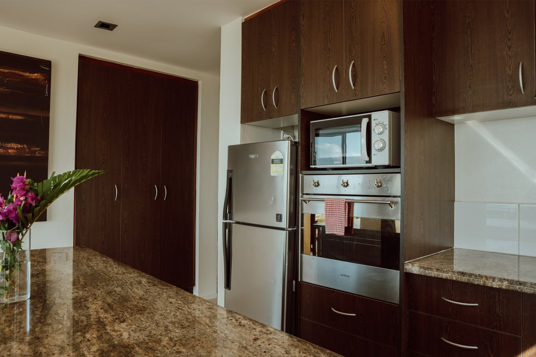 Kitchen with wooden cabinets, stainless steel refrigerator, microwave, oven, and granite countertop. A vase with pink flowers is on the counter.