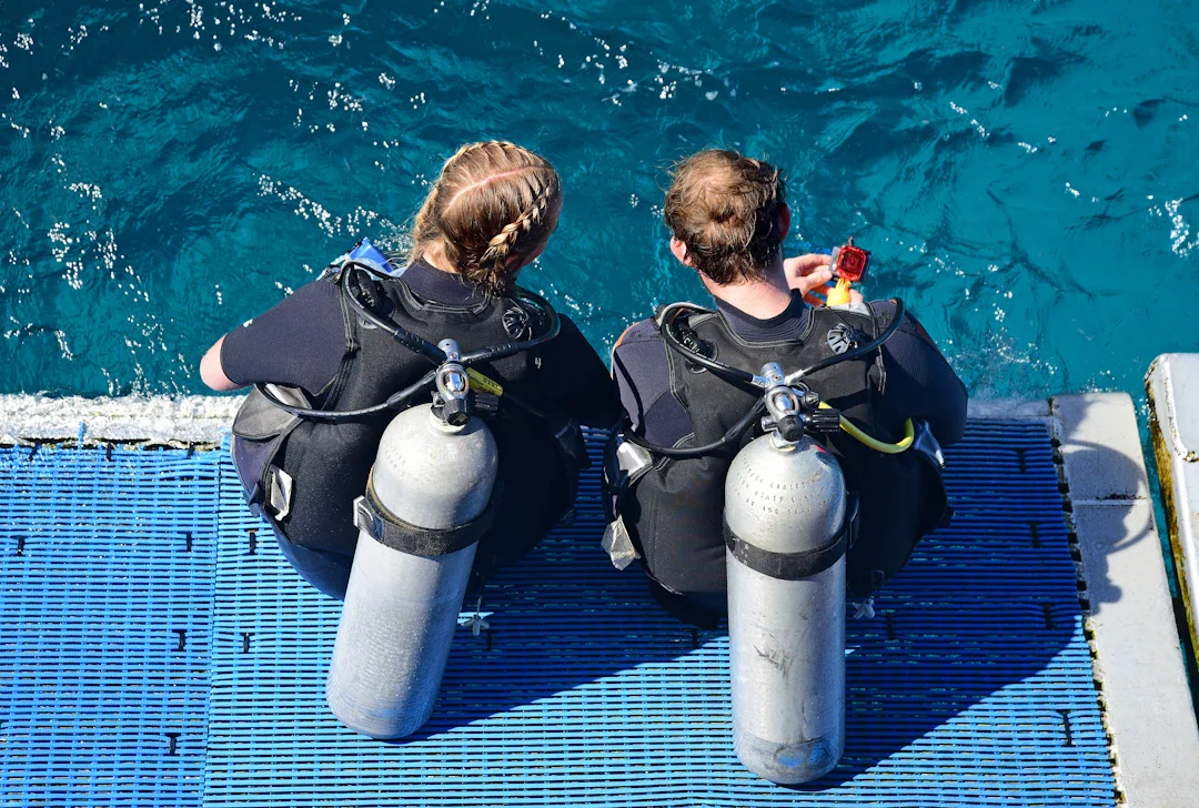 Two scuba divers sitting on a dock, preparing to dive into the water, with their tanks and gear.