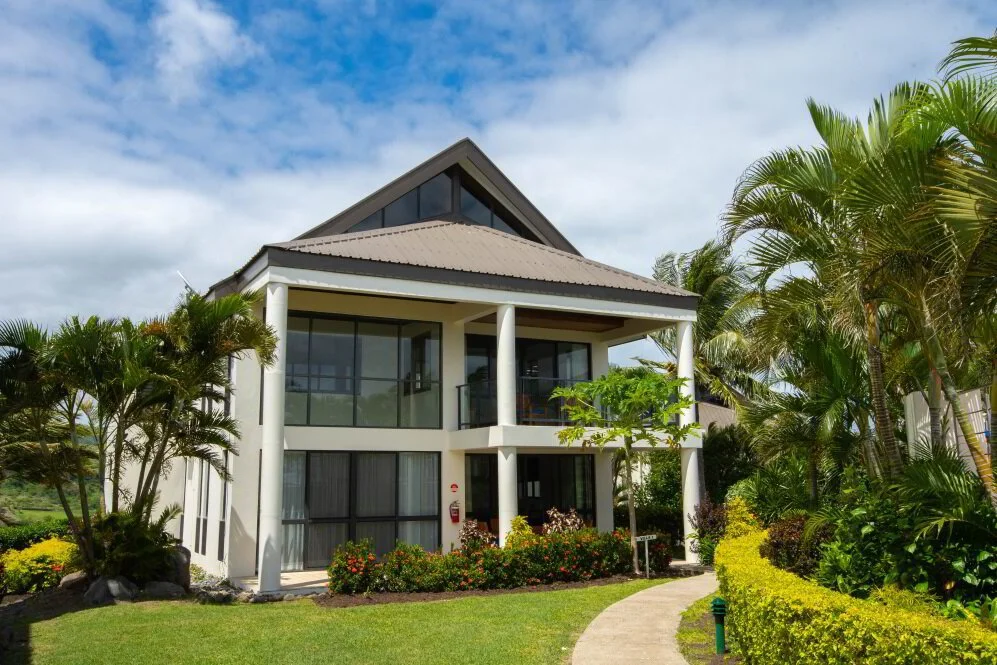 A modern two-story house with large glass windows, surrounded by lush green plants and trees, with a curved pathway leading to the entrance, under a partly cloudy sky.