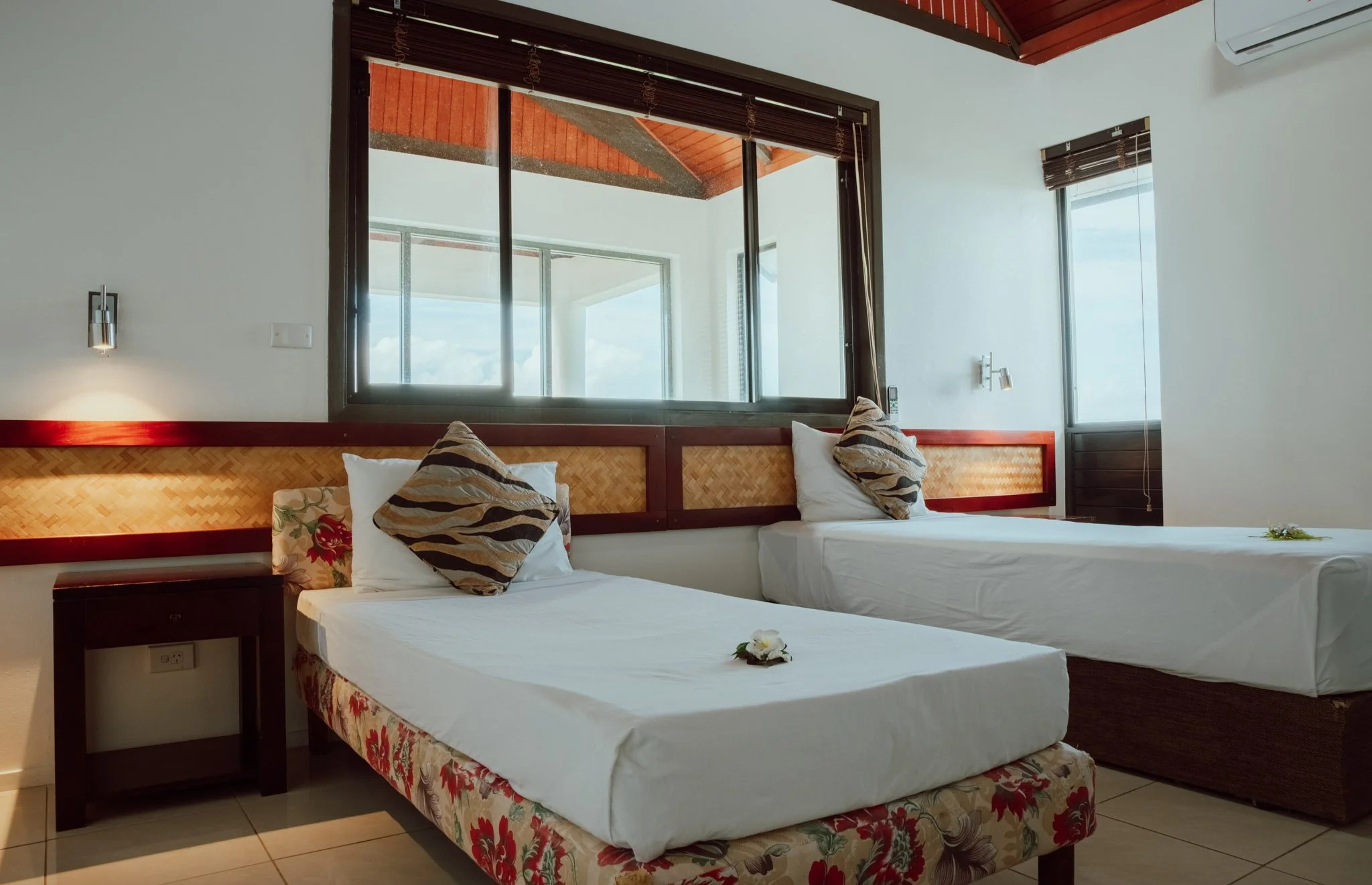 Hotel room with two twin beds, pillows, flowers, large windows, and air conditioning.