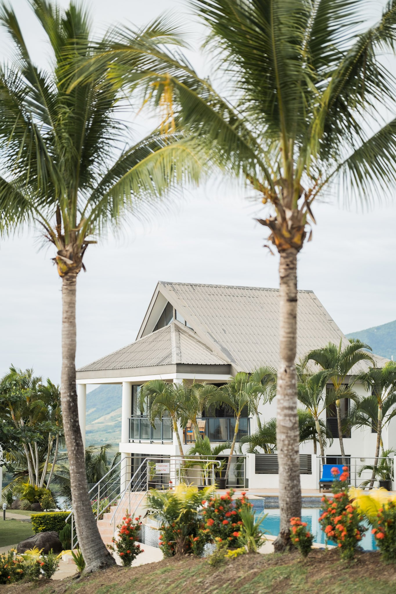 A tropical resort with a building, swimming pool, palm trees, and lush landscaping under a cloudy sky.