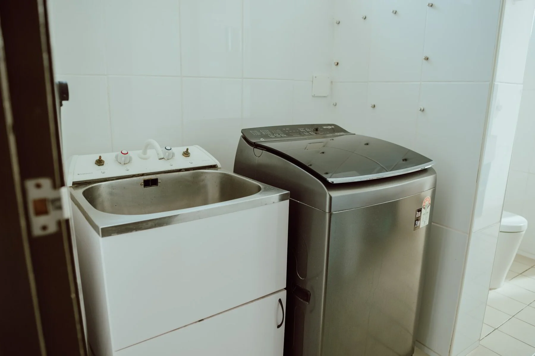 Laundry room with a sink on the left and a top-loading washing machine on the right, against white tiled walls.
