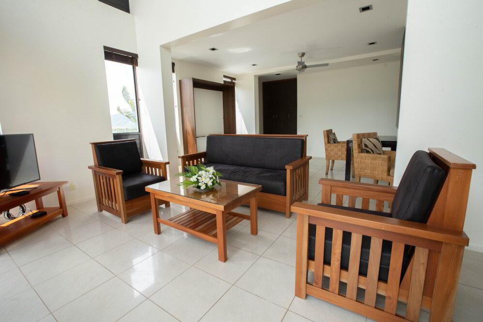 Living room with wooden furniture, black cushioned chairs, a small wooden coffee table with flowers, a TV on a wooden stand, in a bright space with white walls and tiled floor.