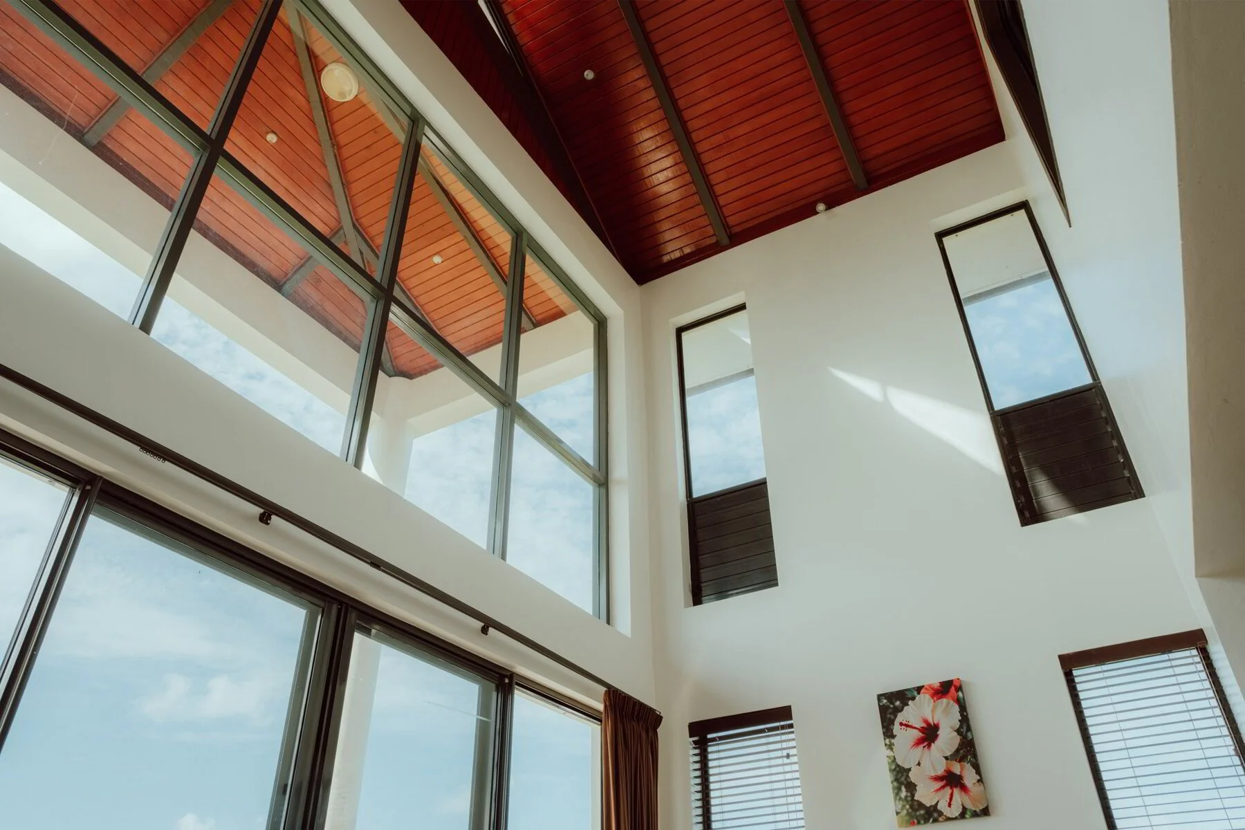 Interior view of a modern house with large glass windows, a wooden ceiling, and a high ceiling with two tilted windows and a flower painting on the wall.