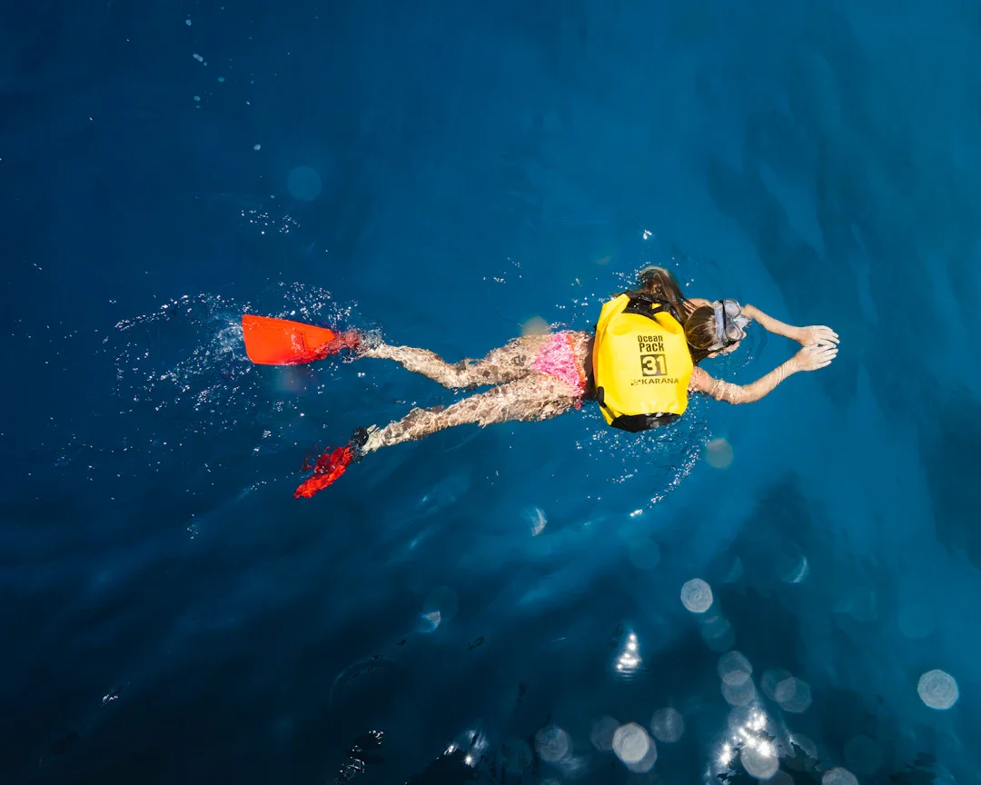 Child snorkeling in deep blue ocean, wearing a yellow life vest labeled 'Ocean Pack 31', pink swimsuit, snorkel mask, and red fins, swimming with outstretched arms.