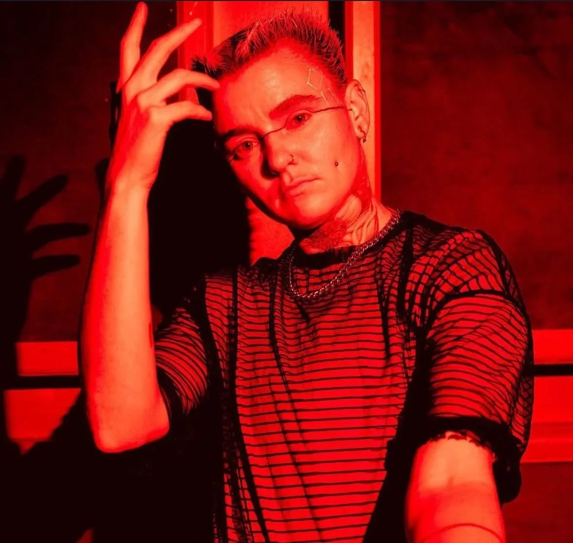 A person with short hair, wearing earrings, a nose ring, a chain necklace, and a sheer black striped shirt, posing in front of a red-lit background.