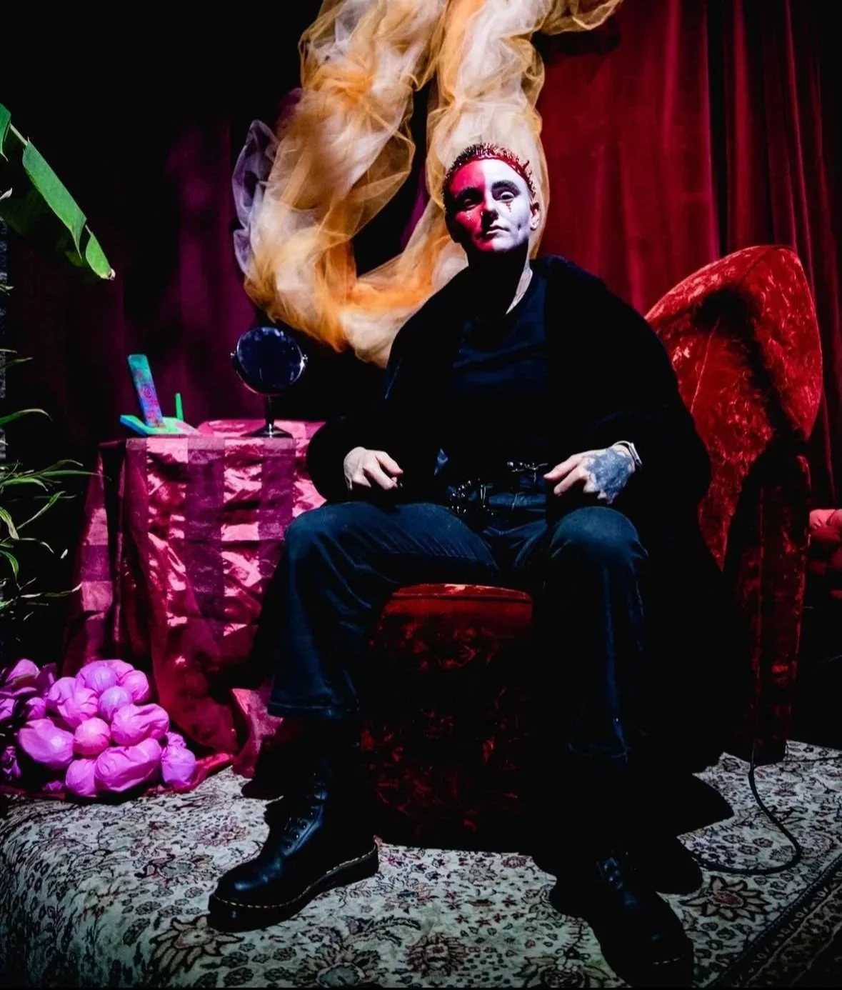 Person with bold makeup sitting on a vintage red velvet armchair in a colorful, theatrical setting with pink and purple decorations and a red curtain in the background.
