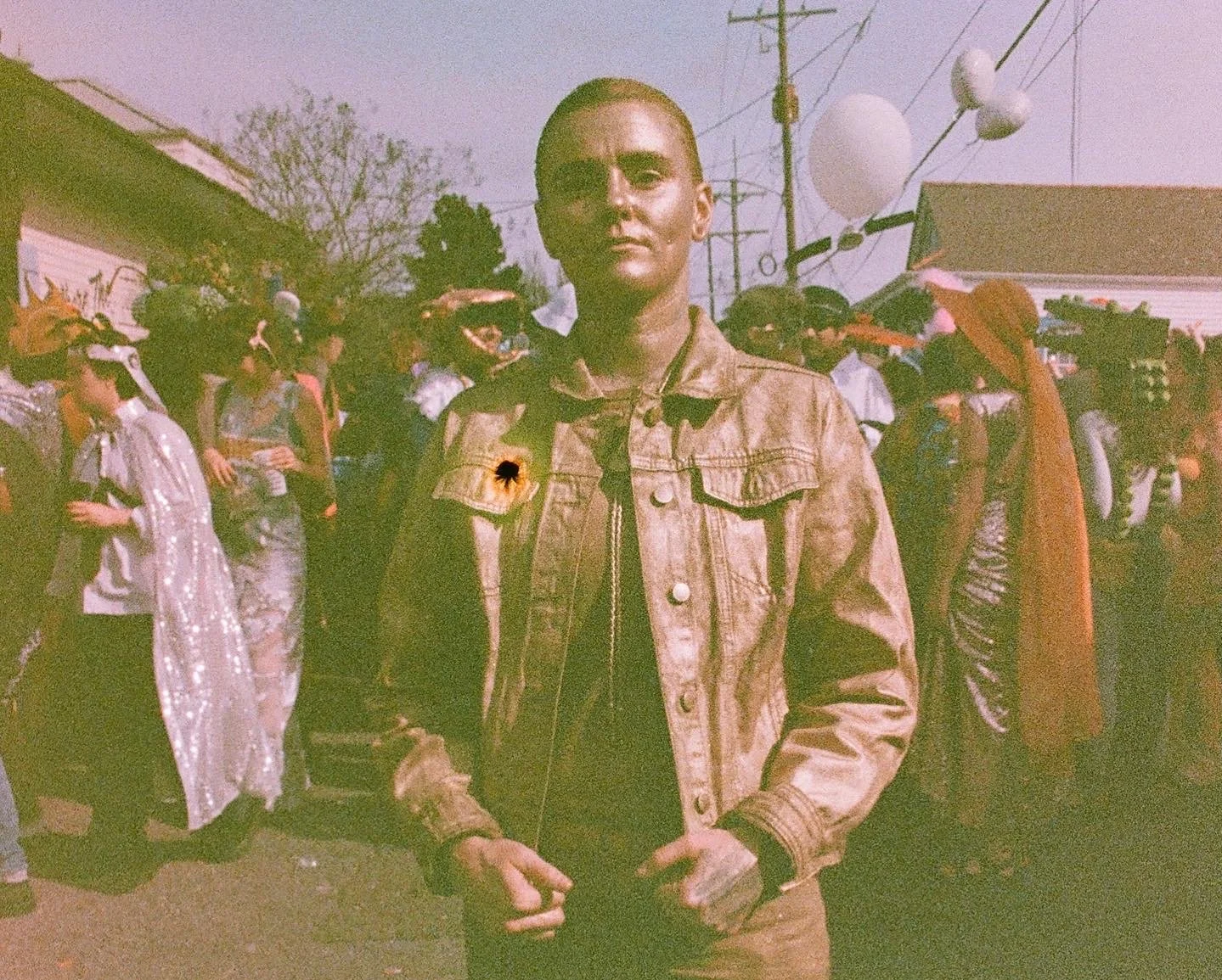 A young person with a shaved head wearing a gold jacket standing outdoors during daytime. There is a crowd of people behind them, some with umbrellas, and houses and power lines in the background.