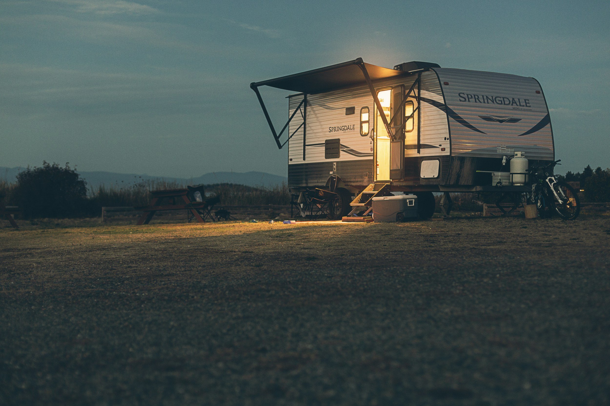 A travel trailer parked on a grassy field during dusk with chairs, a cooler, and a bicycle nearby.
