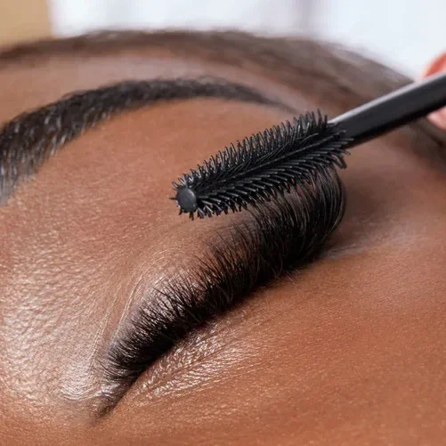Close-up of a person's closed eye with eyelash extensions, with a mascara spoolie brush applying mascara to the lashes.