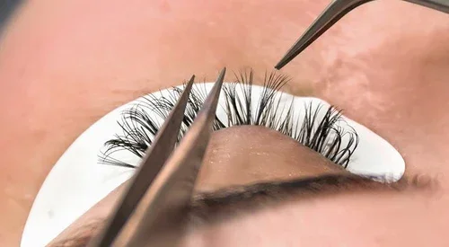 Close-up of a person receiving eyelash extension procedure with tweezers and a patch under the eye.