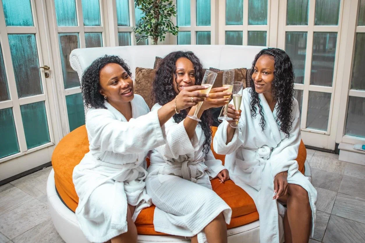 Three women in white bathrobes enjoying drinks and toasts while sitting on a orange cushioned sofa in a bright room with large windows and a plant in the background.