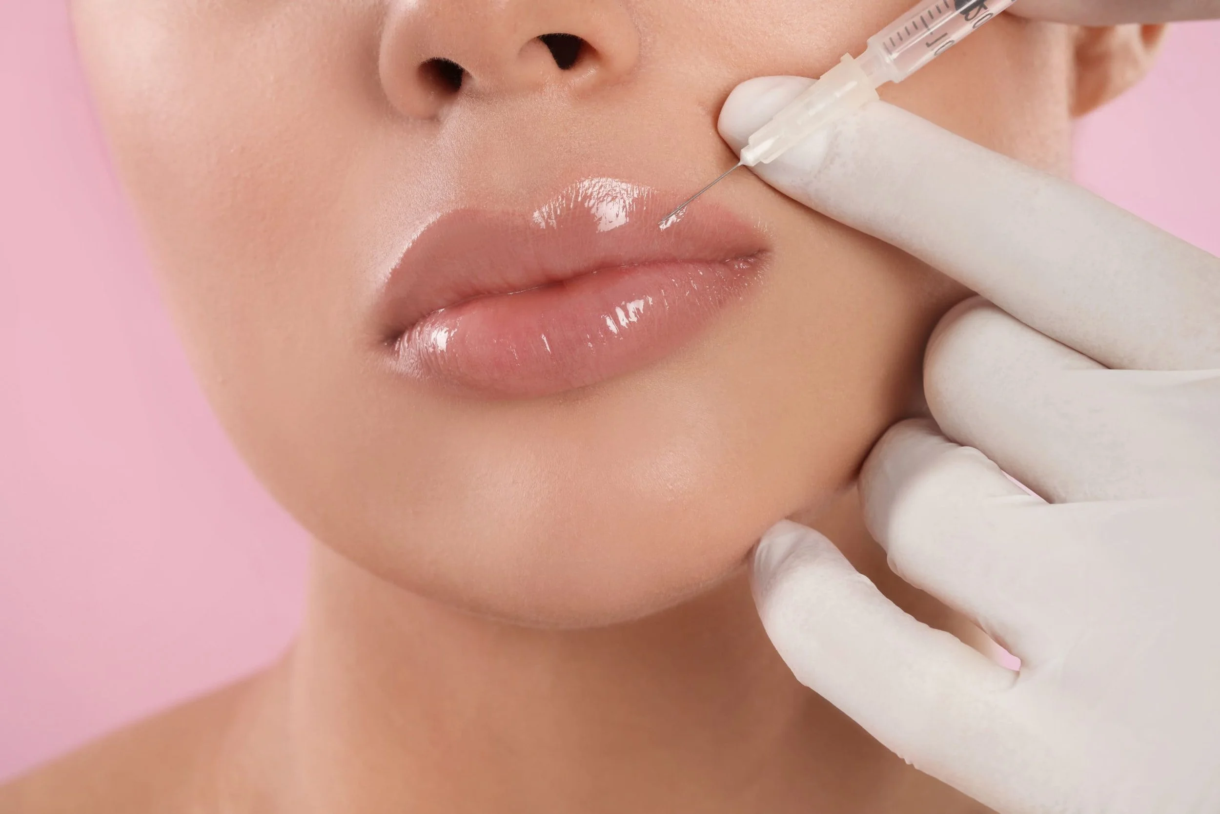 Close-up of a woman's face receiving a lip injection from a healthcare professional wearing gloves, with a pink background.