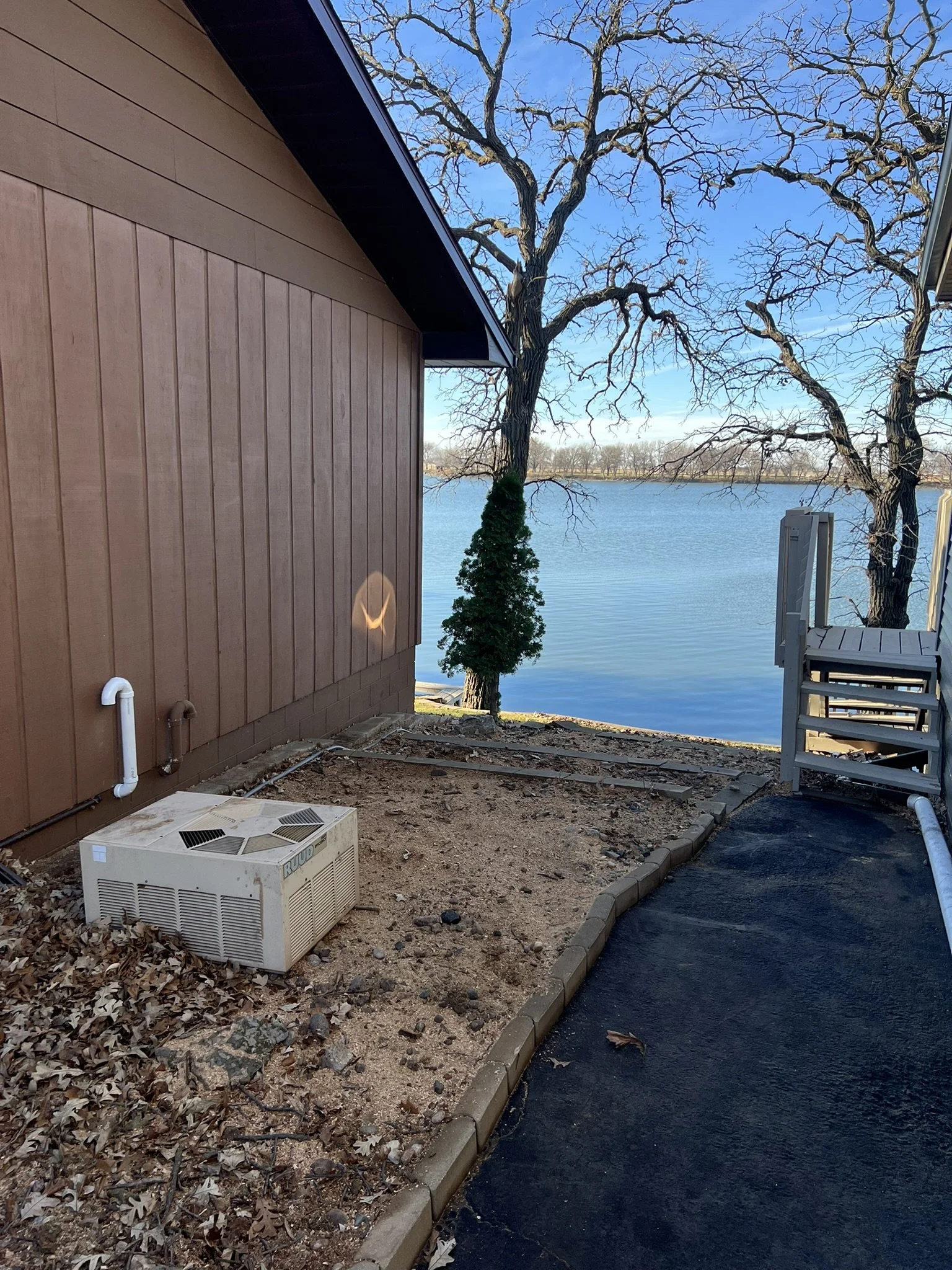 A backyard with a dirt patch beside a brown house, a small tree encased with greenery, and a large leafless tree by a body of water. A staircase leads down to the water, and an air conditioning unit is on the ground.