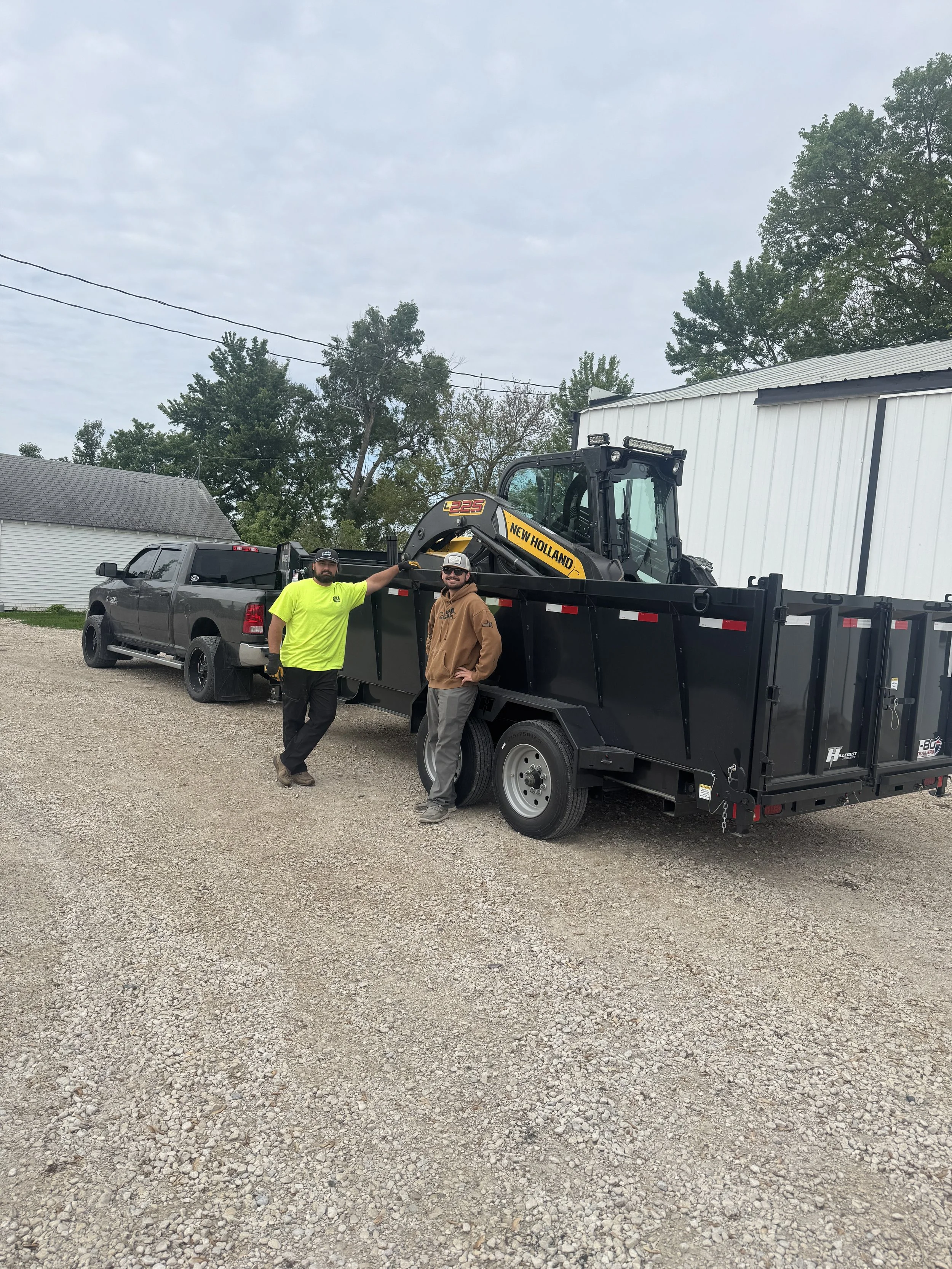 Two men standing next to a black trailer attached to a pickup truck, with a yellow New Holland compact excavator loaded on the trailer, in a gravel lot with trees and buildings in the background.