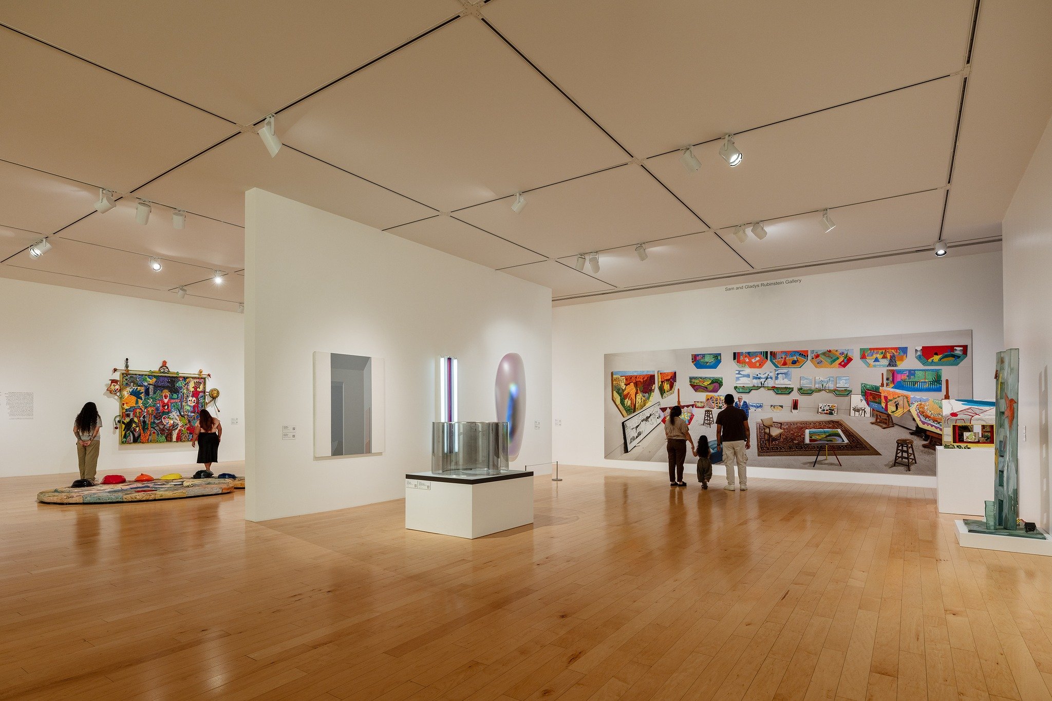 An art gallery with white walls and wooden floors, featuring contemporary paintings and installations. Several visitors are viewing the artwork, with some standing and others sitting on the floor.